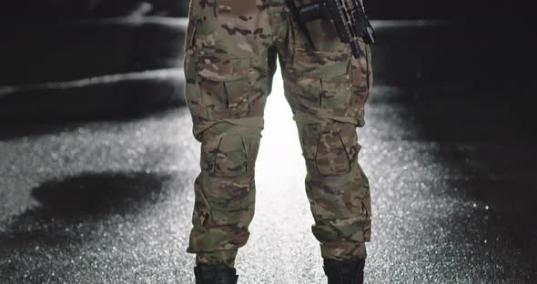 A Soldier in a Special War Uniform Helmet and Gloves Stands in the Dark Leaning Against a Rifle