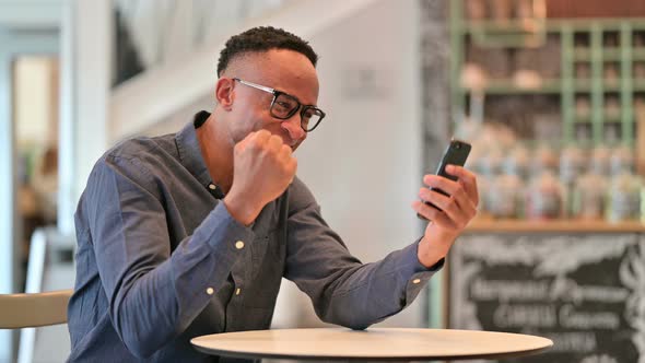 African Man Celebrating Success on Smartphone in Cafe 