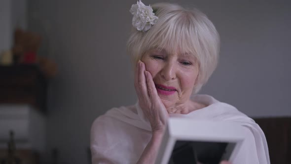 Portrait of Beautiful Excited Caucasian Senior Bride Admiring Old Picture in Photo Frame Sitting in