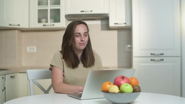 Smiling young woman freelancer working on a laptop in the kitchen from home. Female freelancer