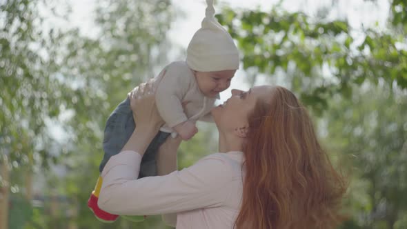 Side View of Beautiful Red-haired Woman Playing with Her Kid in the Park or Garden Close-up