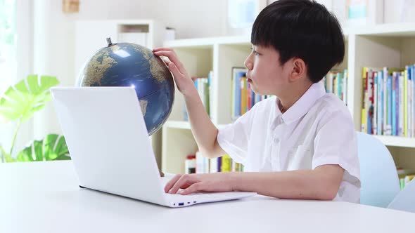 A boy studying while looking at a world map with a globe while looking at a laptop