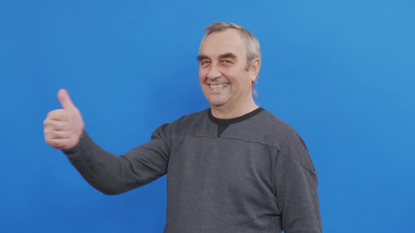 Senior Man Showing His Thumbs Up Approving Your Choice. Studio Shot on Blue Wall.