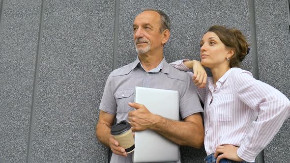 Young Woman and Senior Man are Waiting for a Meeting Standing Near the Grey Wall Outdoors in the