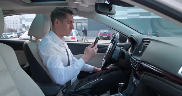 Portrait of a Young Male Entrepreneur Sits in the Driver's Seat in a Car and Uses a Modern