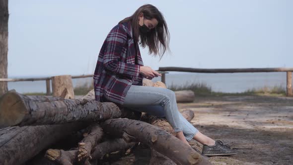 Side View of Brunette Girl in Face Mask Using Phone As Sitting on Log Next To the River. Young