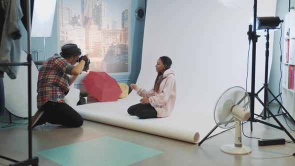 Young Caucasian Photographer Taking Photos of Pretty African Model Sitting on the Floor in Studio