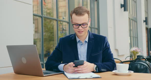 A Businessman Is Sitting at a Table in a Cafe and Texting on His Smartphone