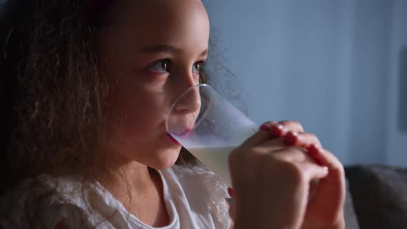 Little Girl Positive Child Drinks Milk While Sitting on Sofa at Home While Watching TV