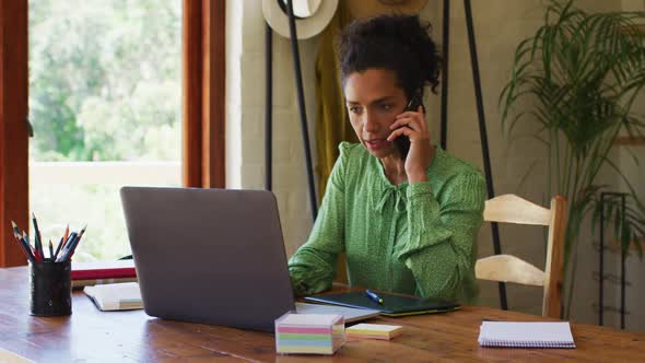 Mixed race woman using laptop and talking on smartphone while working from home