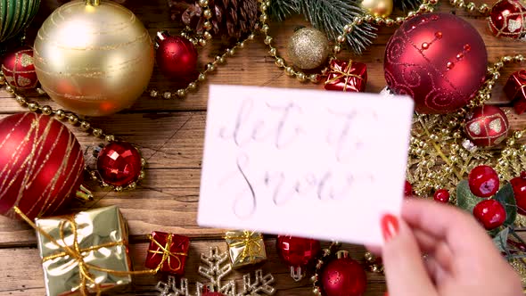 Woman's hand putting a Christmas card with the text LET IT SNOW on a wooden table