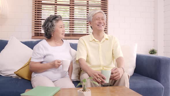 Asian elderly couple drinking warm coffee and talking together in living room at home.