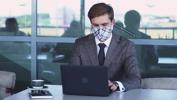 Young Businessman in Medical Masks Is Sitting on the Terrace of a Cafe and Working on a Laptop, View