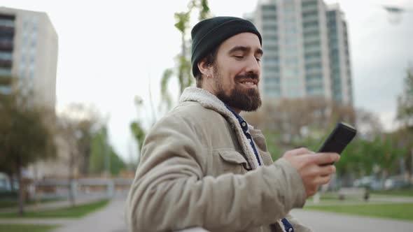 Happy bearded man wearing hat typing by phone