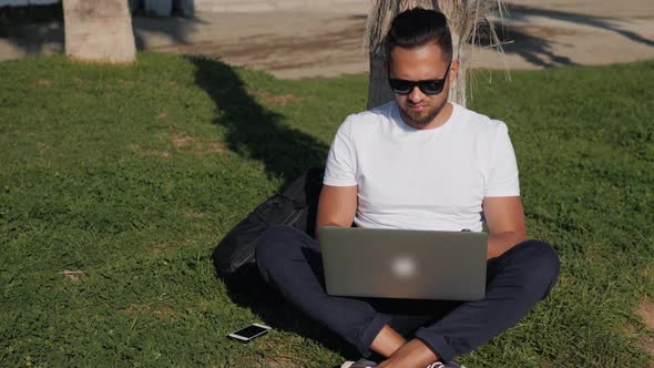 Young Bearded Man Tshirt Wearing Glasses Sitting on the Grass in the Park with His Laptop and