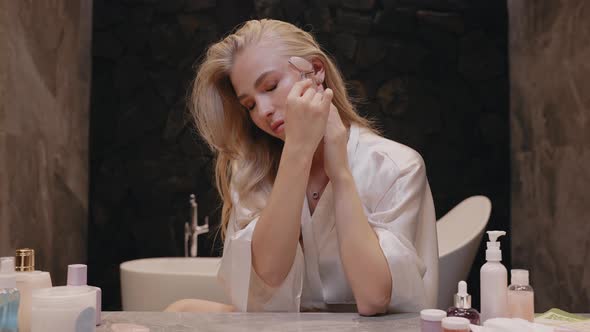 Young Blonde Woman in a White Robe in the Bathroom with a Roller Massager