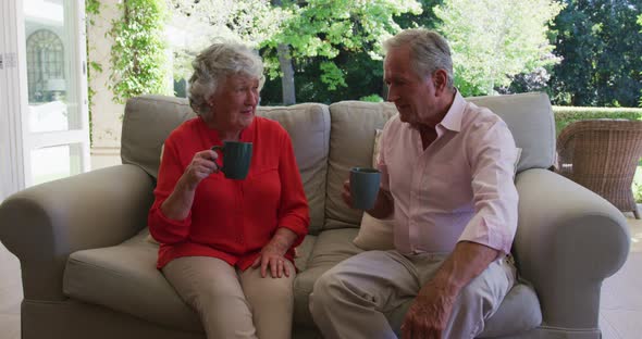Happy caucasian senior couple on couch in sunny living room talking and drinking cups of coffee