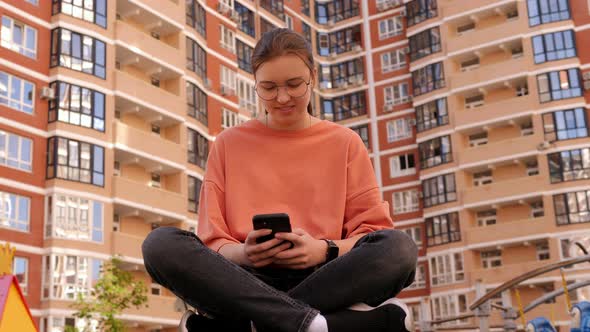 A Teenage Girl is Sitting in the Yard of a Multistorey Building with a Phone