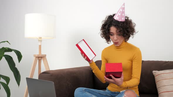 Happy Woman in Party Hat Opens Gift Box and Surprised Sitting in Couch at Home