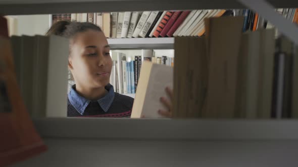 Young African American Student Girl Looking for Neccessary Book on Bookshelf in Big Library