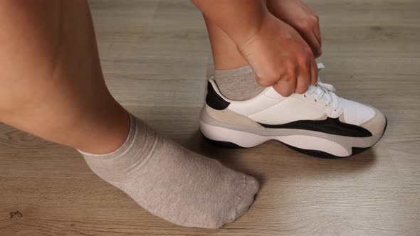 A woman puts a white sneaker on her gray socked foot. Ties her shoelaces