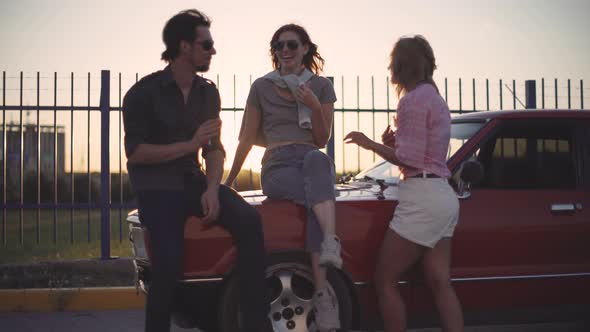 Friends Sit on the Hood of a Car Talk and Laugh at Sunset