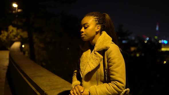 A Young Black Woman is Sad and Shakes Her Head As She Sits in a Park in an Urban Area at Night