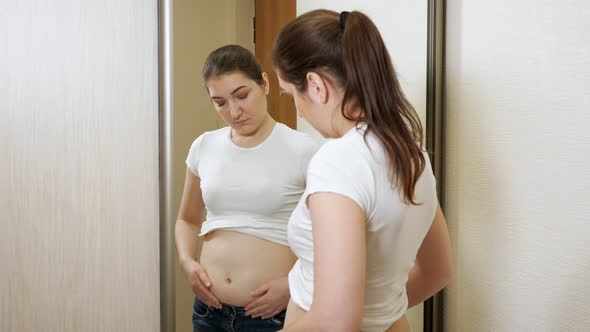 Young Overweight Woman in Front of a Mirror Palpates Her Sides and Stomach