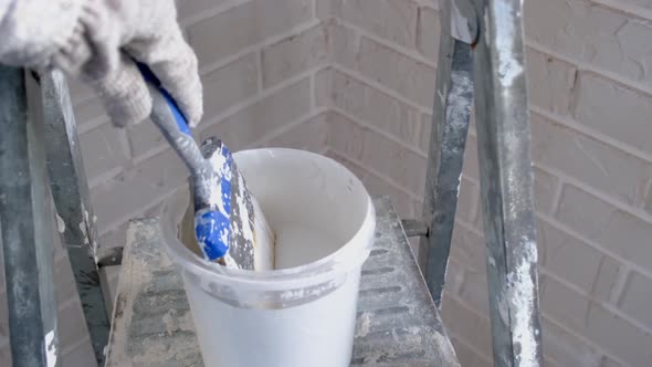 A hand in a work glove dips a paint brush in white paint in a bucket on a ladder against a brick wal