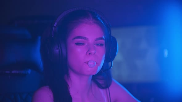 Young Pretty Woman Sitting in Neon Gaming Club and Blowing a Bubblegum - Looking in the Camera