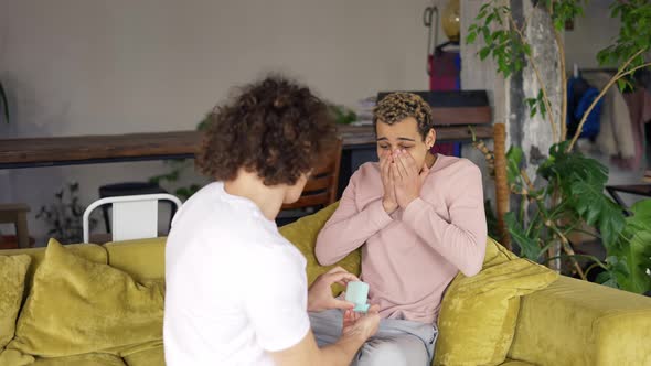 Joyful Man Making Marriage Proposal to His Boyfriend Hugging Together on a Sofa in Living Room