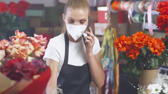 Florist Woman in a Protective Medical Mask Discusses the Order By Talking on the Smartphone
