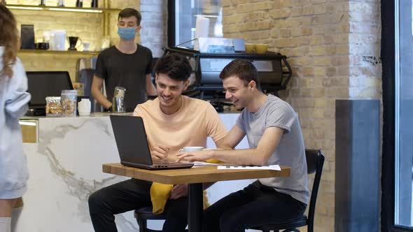 Relaxed Young Colleagues Discussing Startup in Cafe As Male Barista in Face Mask Serving Woman at