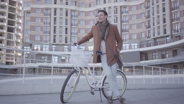 Handsome Serious Man in Brown Coat and Light Blue Jeans Standing with His Bicycle in the City in