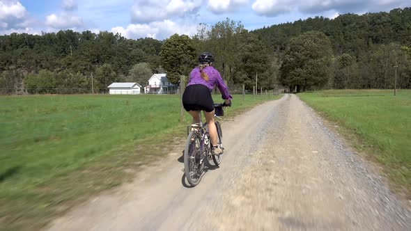 Woman with pony tail on a mountain bike coasting on a gravel road with a farmhouse in the distance.