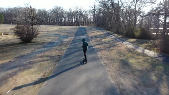 An aerial tracking of a man on an electric skateboard in an empty park on a sunny day. The drone fol