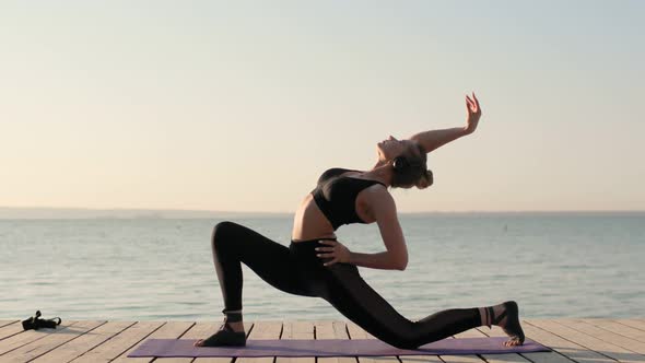 Woman Is Practicing Yoga and Doing Lunge Warrior Yoga Pose on Mat on River Pier.