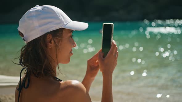 Back View Joyful Young Tanned Woman in Bikini Sits on Sea Beach on Blue Water Background and Uses