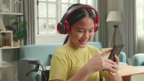 Close Up Of Asian Kid Girl Sitting In A Wheelchair Listening To Music And Using Mobile Phone