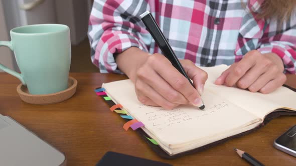 Young Caucasian Attractive Woman Taking Notes in Her Notebook While Sitting in the Kitchen at the