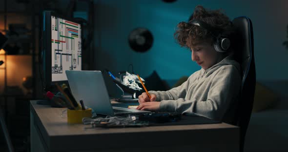 A Boy Sits in Room in the Evening in Front of Computer Screen and Laptop Working on a School Project alt