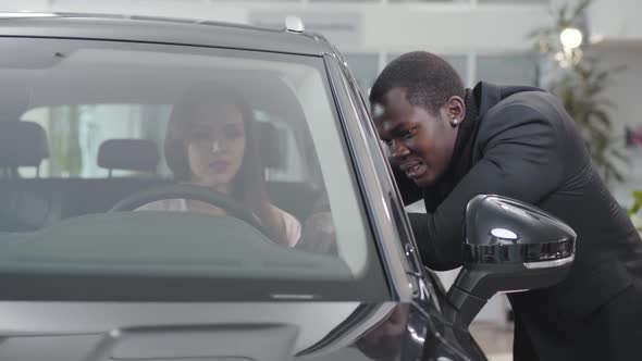 Portrait of Smiling African American Car Dealer Talking To Young Caucasian Woman Sitting in Car