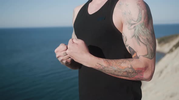 Close Up of Tattooed Man in Tank Top Trains His Arms Outdoors, Sea on Background