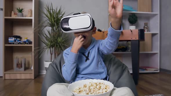 10-Aged Boy in Virtual Reality Headset which Sitting on Comfortable Bean Bag Sofa, Eating Popcorn