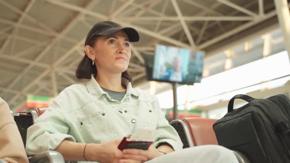 A Woman in a Denim Jacket and Black Baseball Cap Sits with Tickets in Her Hands at the Airport and
