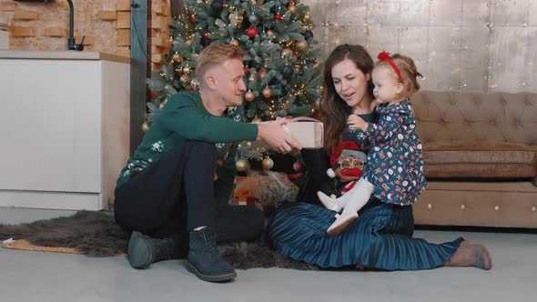 Young Family Near Christmas Tree at Home, Father Gives a Gift Box To His Daughter.