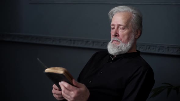 Bearded Senior Aged Man Reading Old Paper Book Sitting in Chair in Dark Living Room Slow Motion