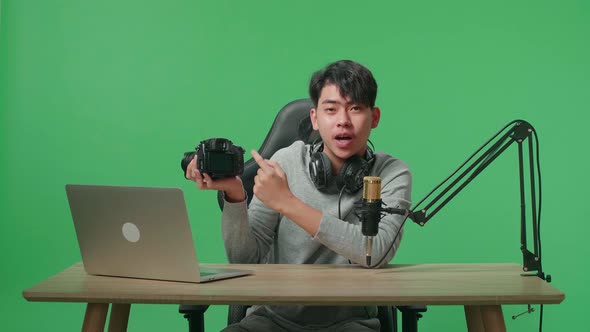 Asian Man With Computer Reviewing Camera While Sitting In Front Of Green Screen Background