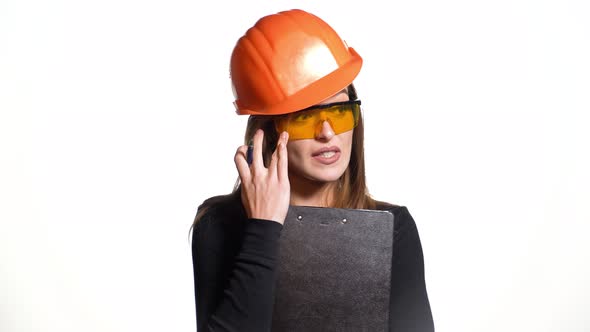 Woman in an Orange Helmet and Protective Eyeglasses is Talking and Holding a Pen and Folder