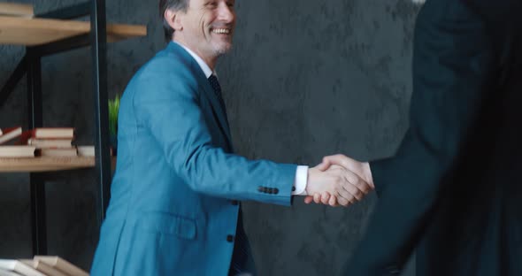 Mature Businessman Shakes Hands with a Colleague Sitting at the Table in the Office